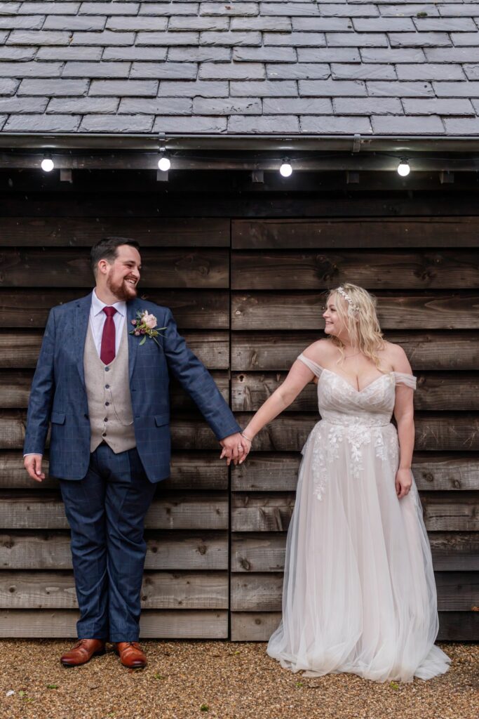 A smiling couple holding hands in front of a rustic wooden backdrop, dressed elegantly for their wedding – Get in Touch with LB Photography for fun and natural wedding photography - connect with fun and friendly wedding photographer
