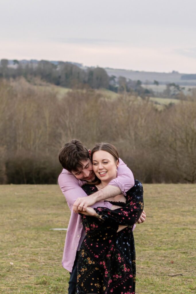 A couple laughing and embracing each other in a grassy field surrounded by trees under a cloudy sky.