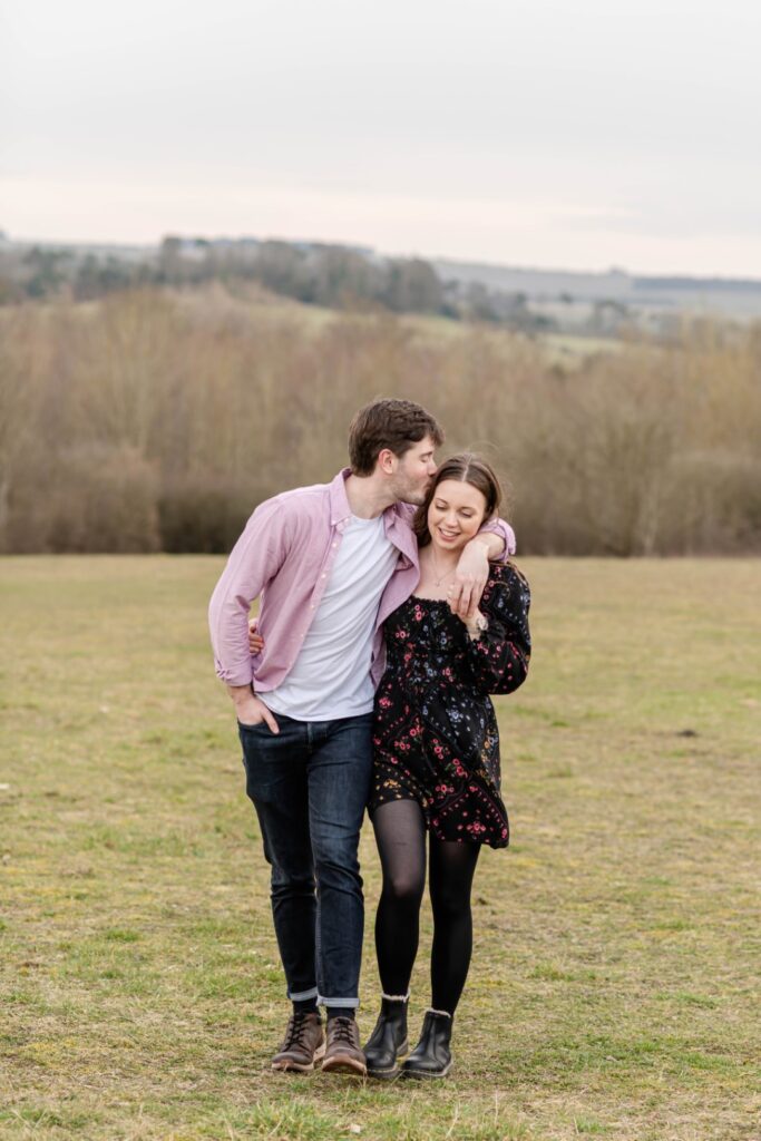 A man and woman share a sweet moment while walking together in a grassy field, surrounded by trees and rolling hills.