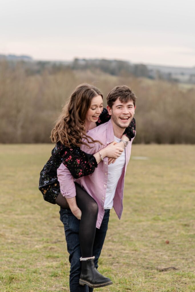 A woman playfully giving a piggyback ride to a smiling man, both enjoying a sunny outdoor setting with trees in the background.
