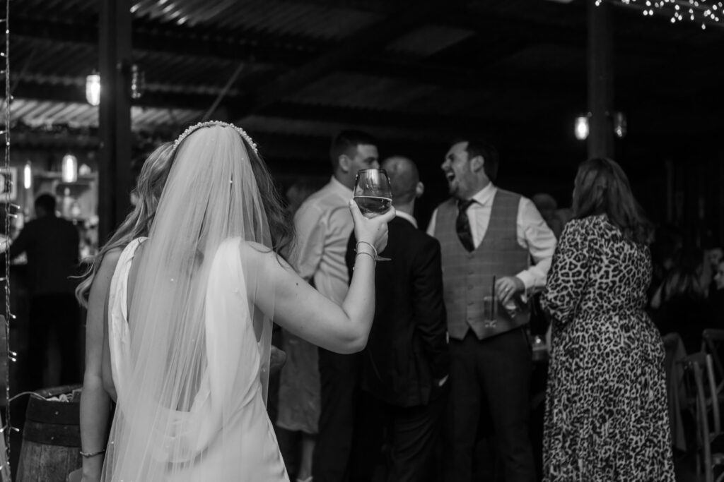 Bride with veil raised a glass during the wedding reception while guests laugh in the background, a candid rainy-day wedding photo by LB Photography.